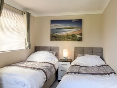 A bedroom with two single beds and a nightstand with a lamp between them and a window with blinds on the left wall at Little Retreat in Liskeard