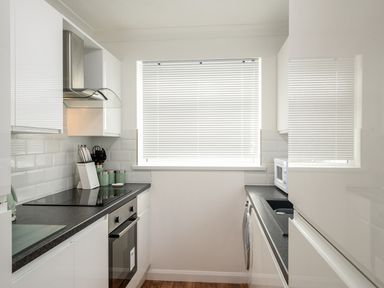 A kitchen with white cabinets black countertops appliances and a window with blinds at Little Retreat in Liskeard