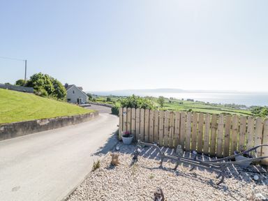 A rural driveway with a gray house and a gravel garden area with decorative figurines near a wooden fence at Tullyally in Redcastle