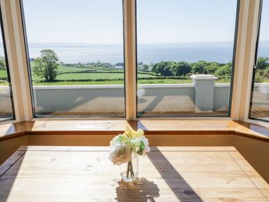 A wooden table with a small flower vase in front of large windows overlooking green fields and a body of water at Tullyally in Redcastle