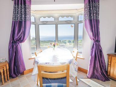 A dining area with a table covered by a tablecloth and wooden chairs by a large window with purple curtains at Tullyally in Redcastle