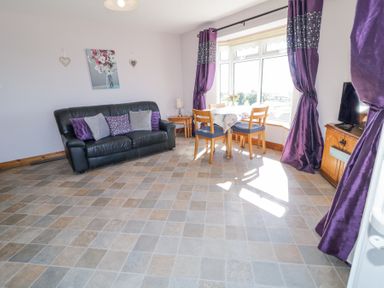 A sitting area with a black leather sofa and purple cushions beside a dining table with wooden chairs near a window with purple curtains and a television on a wooden cabinet at Tullyally in Redcastle