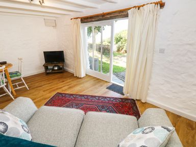 A living room with a gray sofa and patterned cushions a red and gray rug a small TV on a wooden stand and sliding glass doors with cream curtains at The Granary Cottage in Llangrannog near Cardigan