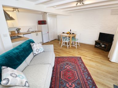 A living room with a grey couch, patterned rug, dining table with chairs, kitchen area, and a television at The Granary Cottage in Llangrannog near Cardigan