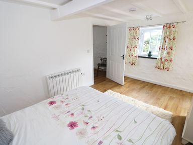 A bedroom with a bed covered in floral bedding a chair in an adjoining room floral curtains on a window and wooden flooring at The Granary Cottage in Llangrannog near Cardigan