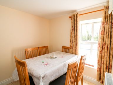 A dining room with a wooden table covered by a floral tablecloth and surrounded by wooden chairs near a window with patterned curtains at Grange Farmhouse in Fethard-on-Sea County Wexford