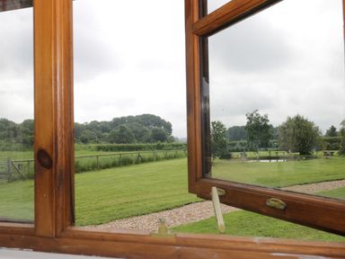 A view of a garden with grass trees and a path visible through a wooden framed open window at The Old Goat Barn at Trout Cottage Somerton