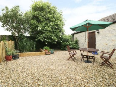 An outdoor patio with gravel ground wooden table and chairs with a green umbrella and plants along a stone wall at The Old Goat Barn at Trout Cottage in Somerton