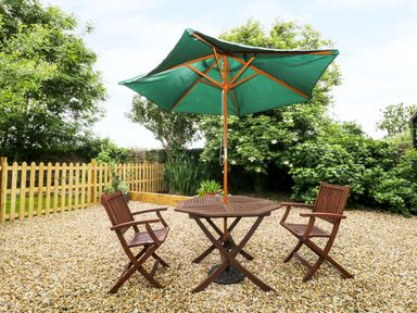A patio area with a wooden table and two chairs under a green umbrella surrounded by trees and a wooden fence at The Old Goat Barn at Trout Cottage in Somerton