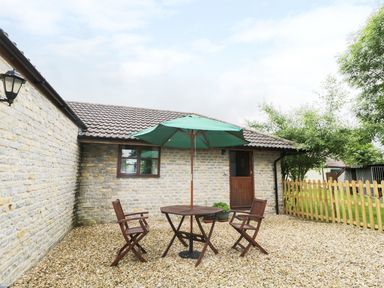 An outdoor patio area with a round wooden table and three wooden chairs under a green umbrella next to a stone wall building and wooden fence at The Old Goat Barn at Trout Cottage in Somerton