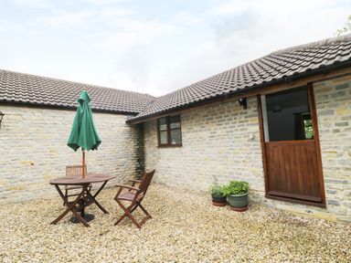 An outdoor patio with a wooden table, two wooden chairs, a folded green umbrella, and two potted plants at The Old Goat Barn at Trout Cottage Somerton