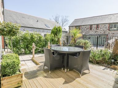 An outdoor patio area with a round glass table and four wicker chairs surrounded by plants and stone buildings at Oh Buoy in Charlestown