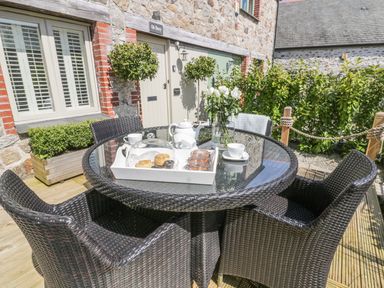 An outdoor patio area with a round glass table and four wicker chairs a tray with teapot scones and cups and a vase with flowers in front of a stone building at Oh Buoy Charlestown