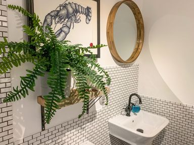 A bathroom with a wall-mounted sink and faucet a round wooden mirror a potted fern on a wooden shelf and a framed lobster illustration on the wall at Oh Buoy in Charlestown