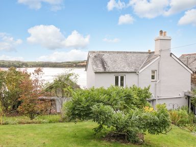 A garden with shrubs and trees behind a white house near a body of water at Clamoak Cottage in Clamoak near Bere Alston
