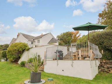 A patio with a wooden bench table and green umbrella surrounded by metal railing near a white house with a garden at Clamoak Cottage in Clamoak near Bere Alston