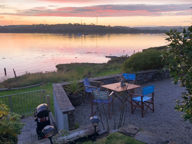 A patio area with four blue chairs and a wooden table overlooking a body of water at sunset at Clamoak Cottage Clamoak near Bere Alston