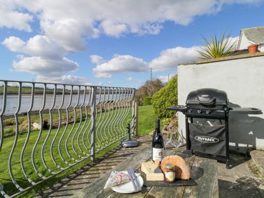 An outdoor patio with a barbecue grill a wooden table with cheese wine and bread near a river at Clamoak Cottage Clamoak near Bere Alston