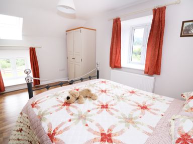A bedroom with a bed covered with a patterned quilt and a teddy bear on it and a wardrobe near windows with red curtains at Cilgwyn Bach in Llandysul