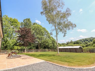 A backyard with lounge chairs on a tiled patio a small tree a larger tree a fence and a shed at Cilgwyn Bach in Llandysul