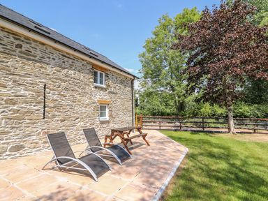 A stone house with two lounge chairs and a wooden picnic table on a tiled patio next to a grassy area with trees and a wooden fence at Cilgwyn Bach in Llandysul