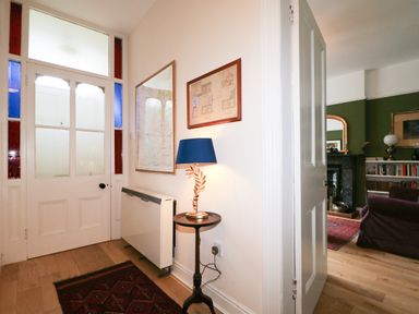 An entryway with a closed white door with stained glass panels a small wooden table with a blue lamp a radiator a framed map and drawing on the wall and a partially open door showing a living room with a purple sofa and bookshelf at Dale House in Lorton