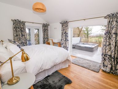 A bedroom with a white bed, gold lamp on a side table, patterned curtains, wooden desk with chair and a hot tub visible outside on a deck at Crooked Hill Cottage in Ammanford