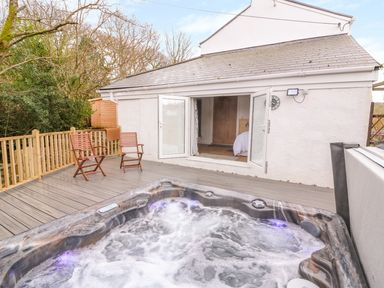 A deck with a hot tub and two wooden chairs outside a white house with open double doors at Crooked Hill Cottage in Ammanford