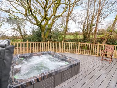 An outdoor wooden deck with a bubbling hot tub and a wooden chair surrounded by trees at Crooked Hill Cottage in Ammanford
