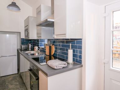 A narrow kitchen with white cabinets blue tiled backsplash and appliances at Crooked Hill Cottage in Ammanford