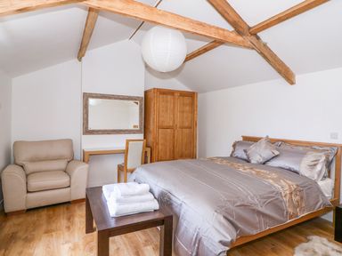 A bedroom with a double bed a wooden wardrobe a beige armchair a desk with a chair a wall mirror and exposed wooden ceiling beams at Crooked Hill Cottage in Ammanford
