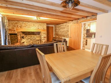 A dining area with wooden table and chairs next to a living room with leather sofas and stone walls at Crooked Hill Cottage in Ammanford