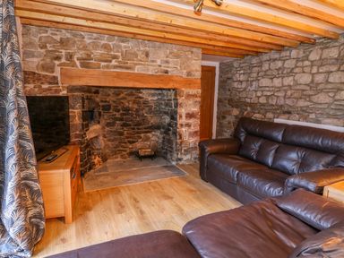 A living room with stone walls wooden beams a stone fireplace brown leather sofas and a wooden TV stand at Crooked Hill Cottage in Ammanford
