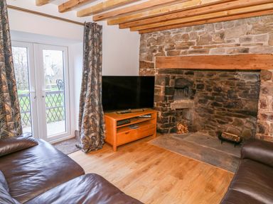 A living room with a stone fireplace two leather sofas a wooden TV stand with a television and glass doors with curtains at Crooked Hill Cottage in Ammanford