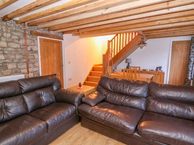 A living room with two brown leather sofas a wooden dining table under stairs and wooden beams on the ceiling at Crooked Hill Cottage in Ammanford