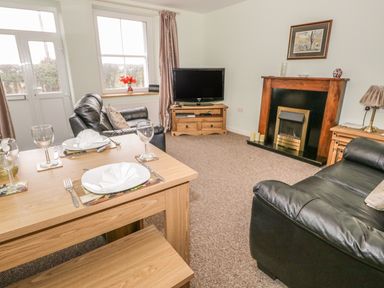A living room with a wooden dining table set for two black leather sofas a wooden fireplace and a television at 2 Park Nook Close in Waberthwaite