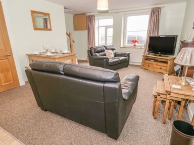 A living room with black leather sofas a wooden TV stand with a television a dining table set and a side table with a lamp at 2 Park Nook Close in Waberthwaite