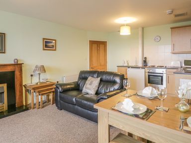 A living room with a black leather sofa a wooden dining table set for two a fireplace and a kitchen area at 2 Park Nook Close in Waberthwaite