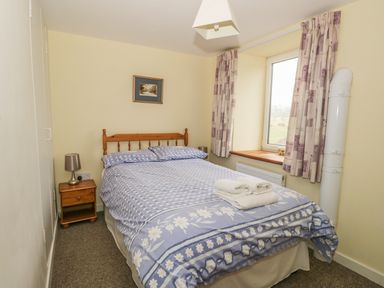 A bedroom with a bed covered in blue patterned bedding a wooden bedside table with a lamp and a window with curtains at 2 Park Nook Close in Waberthwaite