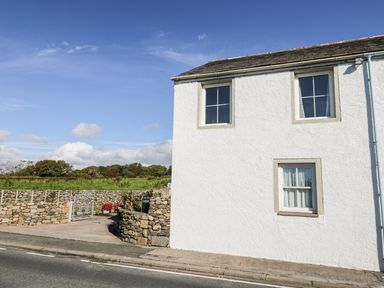 The exterior side view of a white house with three windows next to a stone wall and gated driveway at 1 Park Nook Close in Waberthwaite