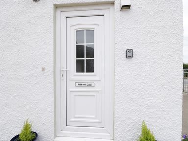 A white front door with a window and sign reading Park Nook Close on a textured white wall with two small plants in pots on either side at 1 Park Nook Close in Waberthwaite