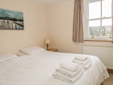 A bedroom with a double bed white bedding folded towels on the bed a window with curtains and a bedside table with lamp at 1 Park Nook Close Waberthwaite