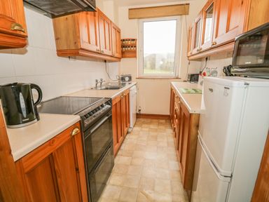 A galley kitchen with wooden cabinets a black stove a white fridge and a window at 1 Park Nook Close in Waberthwaite