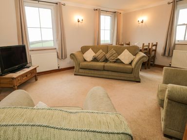 A living room with green sofas a wooden TV stand with a television and a dining area at 1 Park Nook Close in Waberthwaite