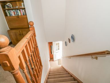 A staircase with wooden handrails and banister leading down to an open door and a wooden cabinet with books on shelves at 1 Park Nook Close Waberthwaite