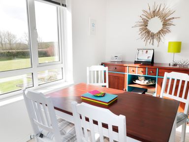 A dining area with a wooden table and white chairs by a window and wooden sideboard with decorative items at Driftwood in Bude