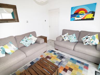 A living room with two gray sofas and patterned cushions a wooden coffee table a colorful rug a wooden framed mirror and a colorful wall painting at Driftwood in Bude