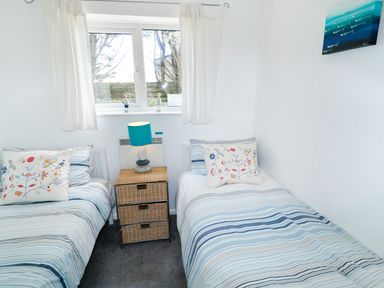 A small bedroom with two single beds with striped bedding a wicker drawer unit with a blue lamp between the beds and a window with white curtains at Driftwood in Bude
