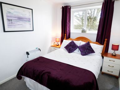 A bedroom with a wooden bed with white and purple bedding two bedside tables with lamps and a window with purple curtains at Driftwood in Bude