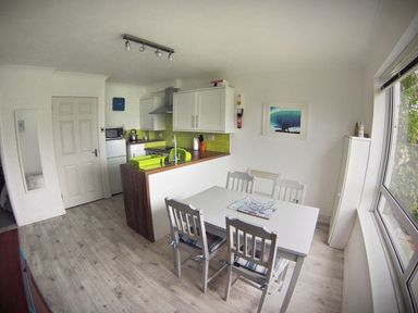 A dining area with a white table and four chairs next to a kitchen with white cabinets and green backsplash at Driftwood in Bude